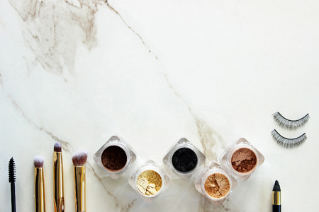 Loose eyeshadow in containers with eyeliner, mascara, and eyelashes with  gold brushes. White marble copy space.の写真素材