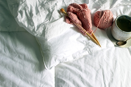 Above view of white bed with pink wool knitting project and a cup of tea.