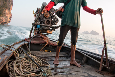 Thai man standing while he drives a wooden long-tail boat at the sea in Ao Nang, Krabi, Thailandの写真素材