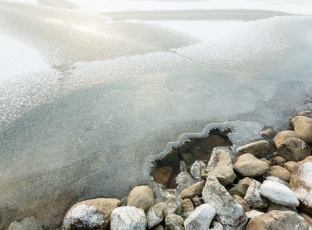 Hole in the ice cover near shore stones at a freezing lake in Finland.