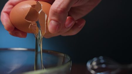 Human hand breaking an egg into a glass bowl. Close-up macro shot against a dark blue backgroundの写真素材
