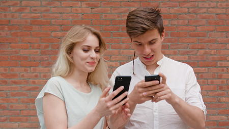 A young blonde couple using smapthones against a brick wall background outdoors. Soft focus. Medium...