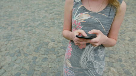 A young lady using a smapthone outdoors against a street paved with cobblestones background. Soft focus close-up shootの写真素材