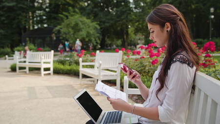 Beautiful brunnette lady sitting on a white bench in the park and using a laptop and a telephone. Medium shotの写真素材