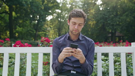 A young man sitting on a white bench in the park and using a phone. Medium shotの写真素材