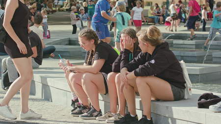 Lublin, Poland - July 2017: Young girls in black clothes are sitting on the edge of a fountain and talkingc covering their eyes from the sun. One stays back to the camera nad swaying hands. Kids are jumping on the fountain in the background. Outdoors. Sunのeditorial素材