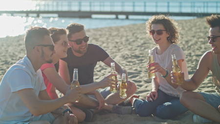 Young people clinking bottles, drinking and having fun on the beach. Medium shot. Soft Focus.の写真素材