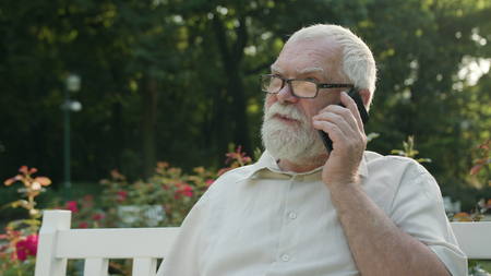 An old man sitting on a white bench in the park and talking on the phone. Medium shot. Soft focus
