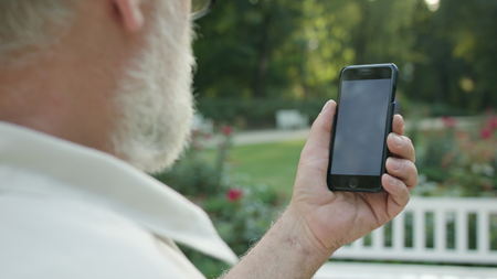 An old man sitting on a white bench in the park and using a mobile phone. Closeup shot. Soft focusの写真素材