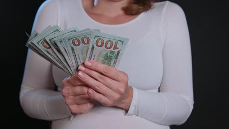 A woman in a white sweater holding cash (US Dollar banknotes) against a black background. Close-up...