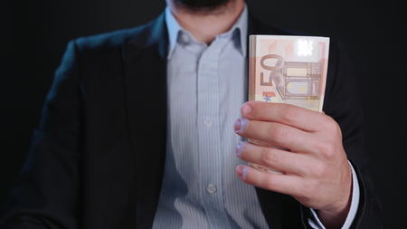 A man wearing a black jacket and a white shirt holding cash (Euros) against a black background....