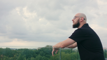 A man with a beard wearing black glasses looks into the distance. Medium shot. Soft focus