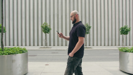 A man with a beard wearing black glasses uses a phone walking in the city street. Long shot. Soft focusの写真素材