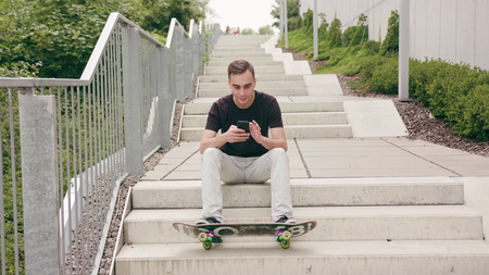 A young man using a phone sitting on the stairs in town. Long shot. Soft focus