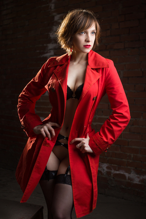 Attractive young woman alluring in sexual lingerie and red coat at grunge industrial setting. Beauty, fashion. Concept: seduction, exhibitionism.の写真素材