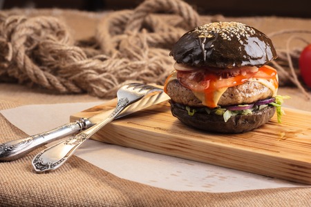 Concept: restaurant menus, healthy eating, homemade, gourmands, gluttony. Trendy glossy burger with beef in black bun with ingredients on messy vintage wooden background.の写真素材