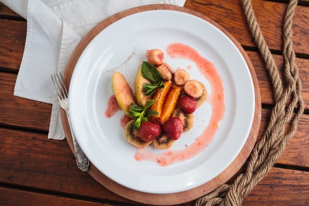 Concept: restaurant menus, healthy eating, homemade, gourmands, gluttony. White plate of fruit flambe. Apple, orange, strawberry, kiwi flambeed in cognac. On weathered wooden table Top-down viewの写真素材