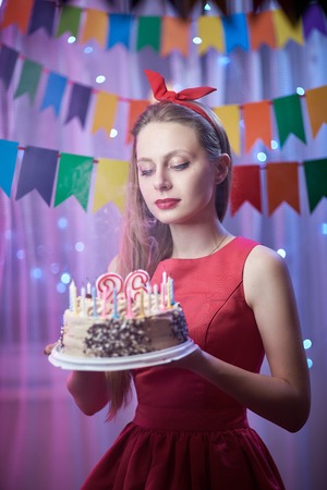 Concept: celebration, birthday. Beautiful young vintage pin up style girl standing in colorful lighted scene holding cake with candles.の写真素材