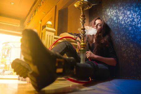 Concept: recreation, millennial, nightlife. Beautiful young woman smoke hookah on floor of empty...