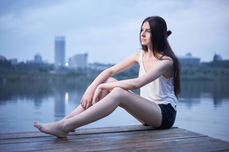 Concept: gritty, millennial. Gorgeous beautiful young woman sunset photoshoot in abandoned urban location near water pier with cityscape on background presenting the contrast of fashion and grunge.の写真素材