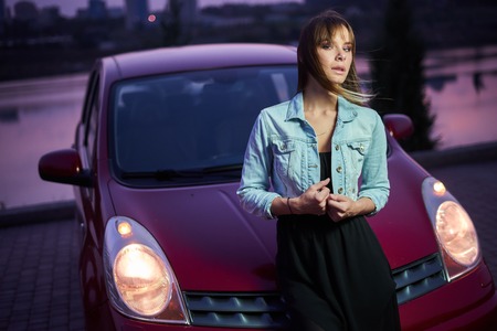 Concept: strong independent young woman with car. Beautiful serious girl fashionably stand near vehicle at twilight pink sunset. Lights are onの写真素材