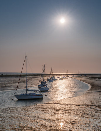 Sunrising over a muddy creek with sailing boats going into the distance in the water at low tideの写真素材