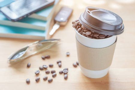 A paper cup of full roasted aroma coffee beans with blurred background of books, smartphone, sunglass and key, good morning concept.の写真素材