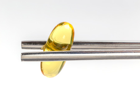 Close up a soft gel capsule is holding by metal chopsticks with white background, essential oil and healthcare concept.の写真素材