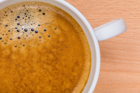 Detail of hot black coffee with foam bubbles in white cup with blurred wooden board in background, morning coffee concept.の写真素材
