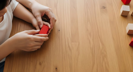 An overhead shot showing an adult's hands gently guiding a child's hands as they hold a red wooden block. The scene takes place on a warm wooden table, conveying themes of learning, guidance, parenting, and developmental play. The composition leaves ample copy space.の素材