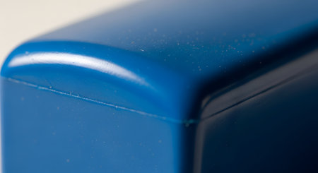 A close-up macro photograph showing the corner of a blue plastic object, possibly a container or case. The image highlights the smooth, curved surface, the seam line, and tiny dust particles, creating an abstract minimalist background. The focus is on the form, texture, and color of the material.の素材