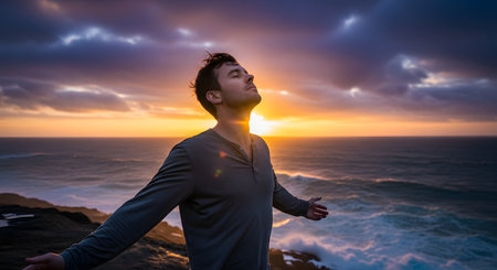 A man stands on a coastal cliff with his arms wide open and eyes closed, embracing the moment as the sun sets over the ocean. His serene expression conveys a profound sense of freedom, peace, spirituality, and connection with nature.の素材