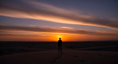 The silhouette of a lone person stands on top of a sand dune, looking out at a breathtaking desert sunset. Footprints trail behind them in the sand, evoking feelings of solitude, adventure, and contemplation.の素材