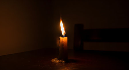 A single candle provides a solitary point of warm light in an otherwise dark room, its flame flickering gently. Melted wax has dripped down its side and pooled at the base on a wooden table, creating a moody and contemplative atmosphere of hope, remembrance, or solitude.の素材
