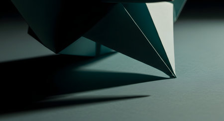 A dramatic, minimalist close-up of a geometric paper sculpture, possibly origami, resting on a dark surface. The sharp angles and clean lines of the folded paper create a strong architectural form, emphasized by the stark lighting and deep shadow.の素材