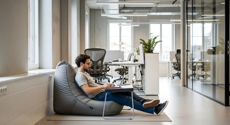 A man casually works on his laptop with a lap desk while sitting comfortably on a grey beanbag chair. This scene takes place in a bright, modern, open-plan office, representing a relaxed and flexible work culture.の素材