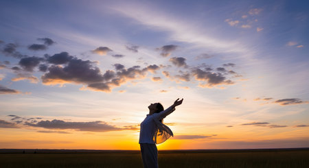 A silhouette of a woman standing in a field with her arms raised in a gesture of freedom and joy against a beautiful sunset sky. This inspiring image represents hope, spirituality, gratitude, and embracing life.の素材