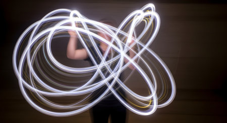 A dynamic long-exposure photograph captures intricate, swirling patterns of white light created by a person in a dark environment. The bright, fluid light trails form a complex, abstract shape, demonstrating the creative technique of light painting. The motion and energy contrast with the dark, still background.の素材