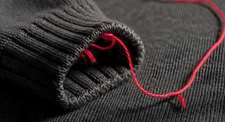 A macro shot focuses on a single loose red thread beginning to unravel from the cuff of a dark grey knitted sweater. The image creates a subtle yet powerful metaphor for a small problem, a flaw, a loose end, or something beginning to fall apart. The contrast between the red thread and the grey fabric draws the viewer's attention.の素材