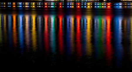 Vibrant, colorful lights from a city skyline or bridge are reflected on the dark, rippling surface of water at night. The long, distorted reflections of red, blue, green, and yellow create a beautiful and abstract pattern.の素材