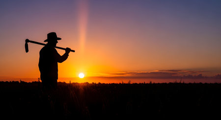 A poignant silhouette of a farmer wearing a hat and holding a hoe over his shoulder as he stands in a field at sunset. The warm, golden sun sinks below the horizon, casting a beautiful glow and creating a long shadow. This image represents the hard work, dedication, and timeless nature of agriculture.の素材