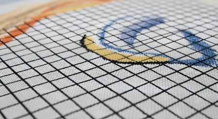 A macro, angled view of a cross-stitch project in progress on white Aida cloth with a printed black grid. Colorful threads in yellow, blue, and black form the beginning of a pattern. The image highlights the texture of the fabric and the precision of the needlecraft hobby.の素材