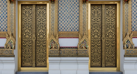 Two magnificent, ornate golden doors of a traditional Thai temple, or Wat, are pictured. The doors and surrounding walls are covered in intricate golden Lai Thai patterns against black and blue backgrounds, showcasing exquisite Thai craftsmanship and architecture.の素材