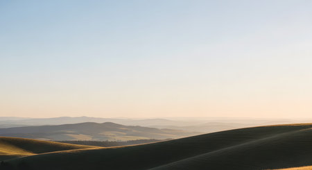 A tranquil landscape photograph of rolling hills bathed in the warm, golden light of sunrise or sunset. The soft contours of the hills create layers that fade into the hazy distance under a clear, pale sky. This serene and peaceful scene evokes a sense of calm and natural beauty.の素材