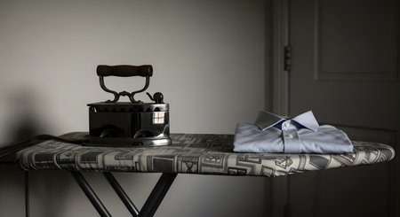 A vintage charcoal iron sits on an ironing board next to a neatly folded, classic striped dress shirt. The scene has a nostalgic and retro feel, contrasting old technology with timeless domestic chores. The moody lighting adds to the classic atmosphere.の素材