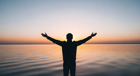 The silhouette of a man standing with his arms wide open, facing a tranquil sea during a beautiful sunset. The scene evokes a sense of freedom, gratitude, spirituality, and connection with nature.の素材