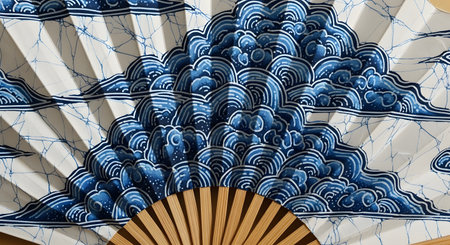 A close-up of a traditional Japanese or Asian folding fan made of paper and bamboo. The fan is decorated with a beautiful blue and white batik-style pattern depicting stylized ocean waves or clouds. This image represents Asian culture, tradition, art, and elegance.の素材