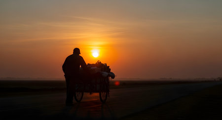 The silhouette of a street vendor pushing his cart along an empty road against a vast, hazy orange sky during sunrise or sunset. The poignant image evokes themes of hard work, perseverance, poverty, and the daily journey of a laborer.の素材