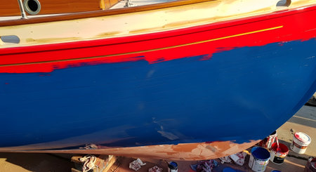 The hull of a wooden boat is being painted with fresh blue paint below a red accent stripe during its maintenance in a dry dock or shipyard. Paint cans and rags are visible on the ground, indicating an ongoing boat repair or restoration project.の素材