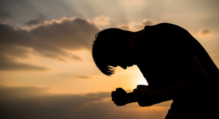A powerful silhouette of a person in despair, bowing their head with clenched fists against a dramatic sunset sky. The image conveys strong emotions of sadness, grief, desperation, struggle, and prayer.の素材